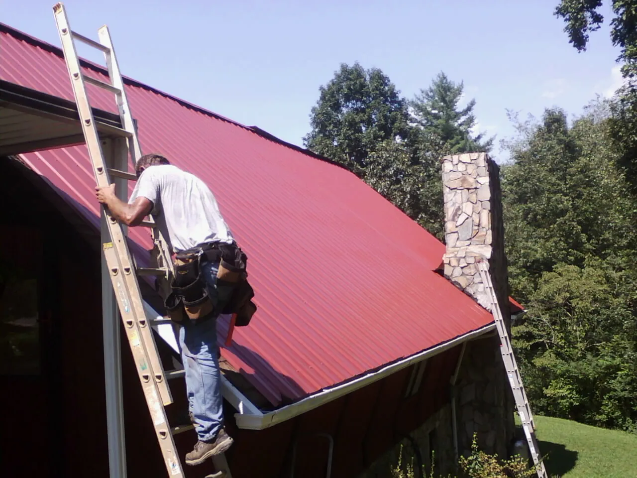 Roofer on a ladder beside a red metal roof and stone chimney — Soffit & Fascia Repair by Roofing Brothers in Cunningham